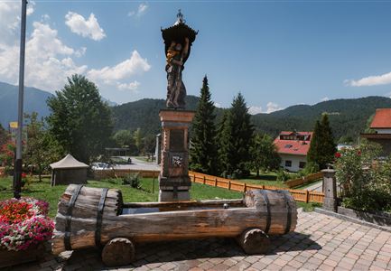 Eine Statue in einer kleinen Stadt, umgeben von Bäumen und einem blauen Himmel. Im Vordergrund steht ein traditioneller Holzofen.