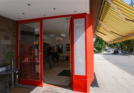 An inviting entrance to a hair salon with a red door. In the background, a few customers and a sunny street can be seen.
