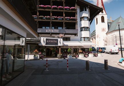 A hotel named "Alpenlove" is located in the foreground. In the background, traditional buildings and a church tower can be seen.