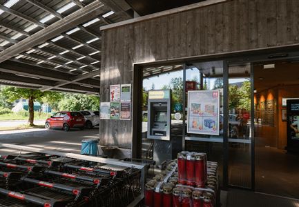 A modern entrance to a shop with large windows and informative posters. In the foreground, there are shopping carts and plenty of daylight is visible.