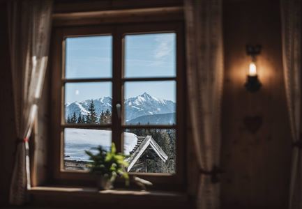 Ein Fenster mit Blick auf schneebedeckte Berge und einen klaren Himmel. Im Vordergrund sind Vorhänge und eine kleine Pflanze zu sehen.