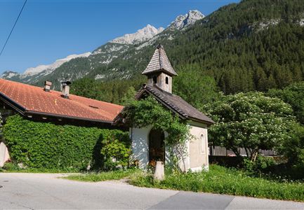 Ein malerisches Dorf mit einem kleinen Kapellengebäude und einer grünen Landschaft. Im Hintergrund sieht man majestätische Berge und blauen Himmel.