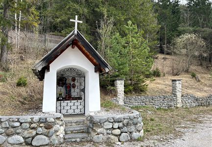 A small chapel with a cross on top stands in nature. It is surrounded by stones and is located in a wooded environment.
