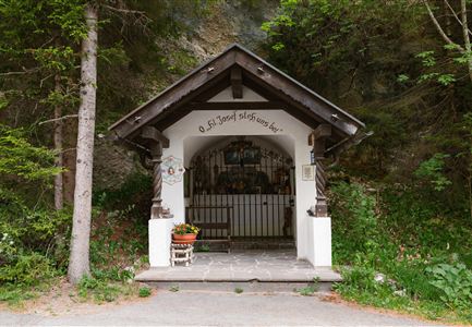 Ein kleiner, einladender Eingang zu einer Kapelle in der Natur. Umgeben von Bäumen und mit einer sitzenden Blume begrüßt er die Besucher.