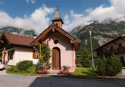 A small chapel with a pointed roof is situated in the midst of mountains. Colorful flowers surround the building and the clouds partially obscure the sky.