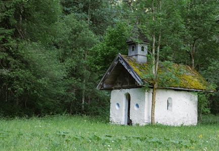 Eine kleine, charmante Kapelle steht inmitten eines grünen Waldes. Die Fassade ist mit Moos bedeckt und umgeben von Bäumen und Wiese.