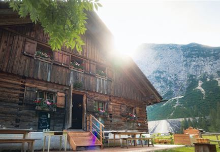 Ein traditionelles Holzhaus in den Bergen mit blühenden Fenstern. Im Hintergrund erheben sich majestätische Berge unter klarem Himmel.