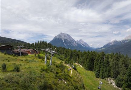Eine malerische Berglandschaft mit grünen Wäldern und einem Skilift. Im Hintergrund sind majestätische Berge und ein klarer Himmel zu sehen.