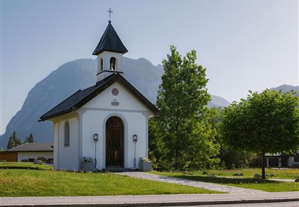 A small, white chapel with a pointed roof stands in the picturesque landscape. In the background, tall mountains and a clear sky are visible.