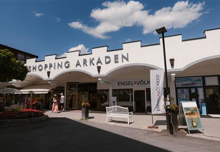 A modern shopping center with the inscription "SHOPPING ARCADES". In the foreground, visitors and a blooming landscape can be seen.
