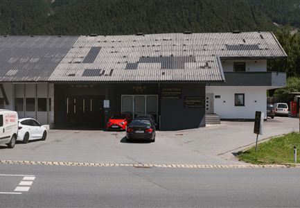 A modern building with an asymmetrical roof and several windows. In front of the building, various cars are parked in an asphalted parking lot.