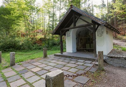 A small chapel surrounded by trees. The entrance is secured with a chain and the path is paved with stones.