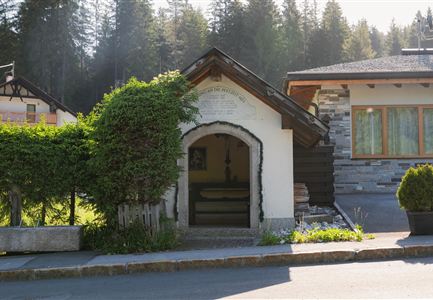 A small, charming building with an arched entrance door, surrounded by lush greenery. In the background, a modern house and tall trees can be seen.