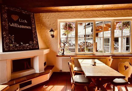 Ein einladender Innenraum mit Holzstühlen und einem Tisch. An der Wand hängt ein Willkommensschild, und durch das Fenster sieht man eine blühende Terrasse.