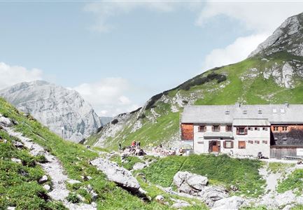 Eine malerische Berghütte umgeben von grünen Wiesen und majestätischen Bergen. Der Himmel ist klar und es herrscht eine ruhige, natürliche Atmosphäre.