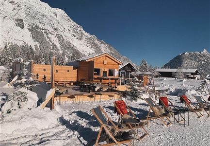 A cozy mountain cabin surrounded by snow and mountains. In the foreground are lounge chairs for relaxing.