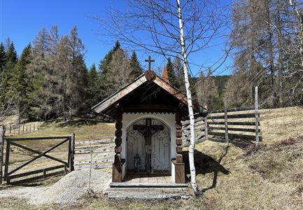A small, decorative chapel made of wood stands in a rural setting. In the background, trees and a fence can be seen under a clear blue sky.