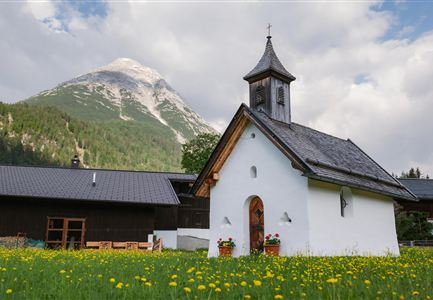 Eine kleine, weiße Kapelle steht inmitten einer blühenden Wiese. Im Hintergrund ragt ein majestätischer Berg unter einem bewölkten Himmel empor.