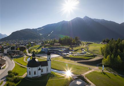 A picturesque landscape with a small church and mountains in the background. The sun shines over the green valley and the surrounding forests.