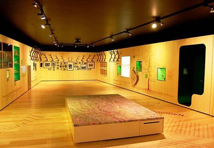 Ein moderner Ausstellungsraum mit hölzernen Wänden und verschiedenen Kunstwerken. In der Mitte befindet sich ein informationstragender Tisch.