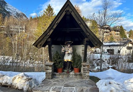 A wooden statue stands under a small roof in a winter landscape. In the background, snow-covered mountains and some houses can be seen.