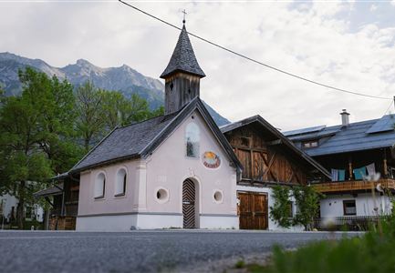 Eine kleine, charmante Kirche mit einem spitzen Turm steht in einer malerischen Umgebung. Im Hintergrund sind Berge und traditionelle Gebäude zu sehen.