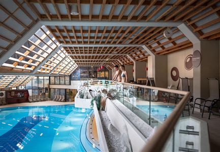 Ein moderner Wellnessbereich mit einer großen Schwimmhalle und einer hellen Holzdecke. Menschen stehen am Rand des Wassers und genießen die entspannende Atmosphäre.
