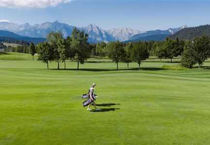 A female golfer walks with her equipment across a green golf course landscape. In the background, mountains and a clear blue sky can be seen.