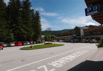 Eine Parkfläche vor einem Gebäude mit vielen Autos. Im Hintergrund sind Bäume und Berge sichtbar.