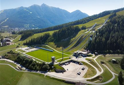 Eine beeindruckende Landschaft mit grünen Wiesen und Bergen im Hintergrund. Im Vordergrund sind Sprungschanzen und Sporteinrichtungen zu sehen.