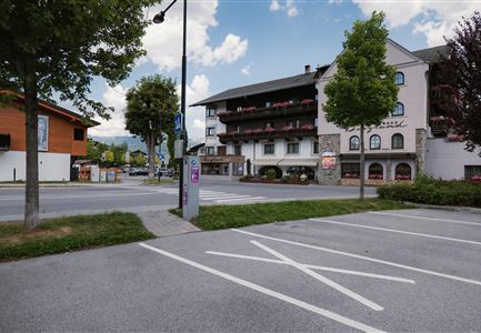 Eine ruhige Straßenszene mit einem Parkplatz und mehreren Gebäuden. Im Hintergrund sind Bäume und ein blauer Himmel zu sehen.
