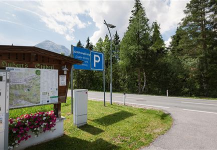 Ein Parkplatz an einer Straße, umgeben von Bäumen und Natur. Ein Informationsschild mit einer Karte ist ebenfalls sichtbar.