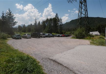 A parking lot in a rural setting with several parked cars. In the background, trees and a mountain landscape are visible.