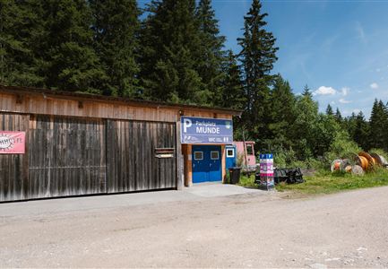 Ein Holzgebäude mit einem Eingang und einem Schild, das "Parkplatz Munde" zeigt. Im Hintergrund sind Bäume und einige Gegenstände zu sehen.