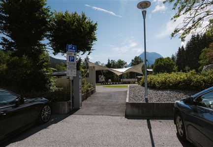Eine moderne Eingangssituation mit zwei Autos und einer begrünten Umgebung. Im Hintergrund sind Bäume und ein Berg zu sehen, unter einem klaren blauen Himmel.