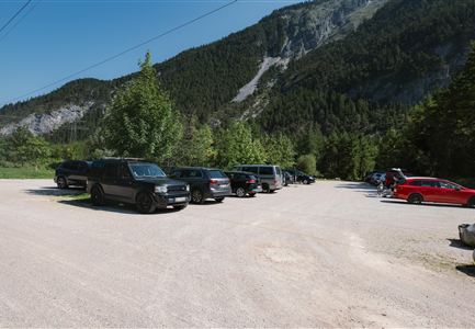 Ein Parkplatz mit mehreren Autos, umgeben von Bäumen und Hügeln. Im Hintergrund sind grüne Berge zu sehen.