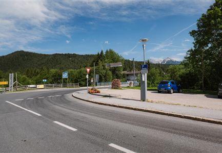 Eine kurvenreiche Straße in einer ländlichen Gegend mit Blick auf Wälder und Berge im Hintergrund. Ein blauer Pkw parkt an einer Parkbucht.
