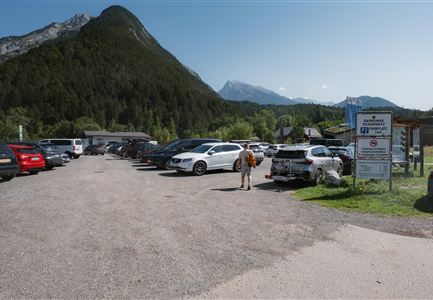 Ein Parkplatz in einer bergigen Umgebung mit vielen Autos. Im Hintergrund sind grüne Bäume und Berge zu sehen.