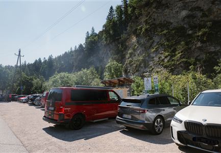 A parking lot with several cars, surrounded by trees and rocks. In the background, gently rolling landscapes are visible.