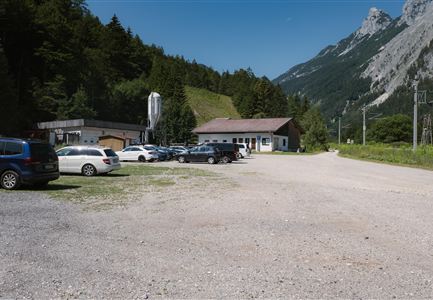 A parking area with several vehicles and a small building in a mountainous setting. In the background, green trees and rocks are visible.