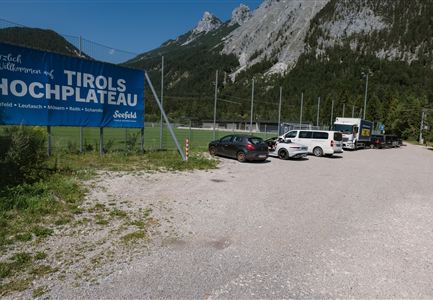 Ein Parkplatz neben einem Weg mit mehreren Autos. Im Hintergrund gibt es eine Berglandschaft und ein Schild, das willkommen heißt im Tiroler Hochplateau.