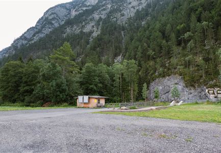 Eine kleine Hütte steht in einer grünen Umgebung mit Bergen im Hintergrund. Der Boden besteht aus Kies und Gras, umgeben von Bäumen.