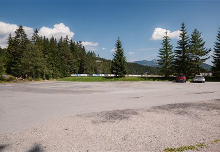 Ein ruhiger Parkplatz umgeben von grünen Bäumen und Bergen im Hintergrund. Der Himmel ist teilweise bewölkt und es sind einige Autos zu sehen.