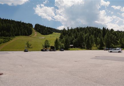 Ein Parkplatz in einem hügeligen Waldgebiet. Im Hintergrund sind Bäume und eine Rodelbahn sichtbar.