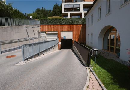 A driveway to an underground garage with a smooth road and a modern building in the background. On the side, there is a green meadow.