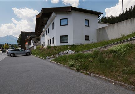 Ein modernes, zweistöckiges Haus steht an einer Straße in den Bergen. Im Vordergrund parkt ein silbernes Auto.