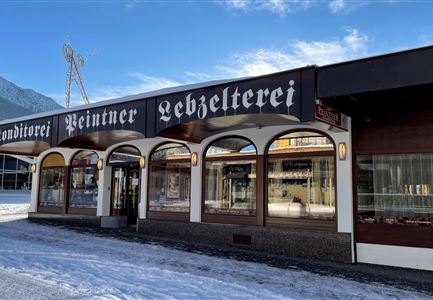 Ein traditionelles Geschäft mit dem Namen "Peintner Lebzelterei" in einer winterlichen Landschaft. Die Fassade hat große Fenster und einen stilvollen Schriftzug.