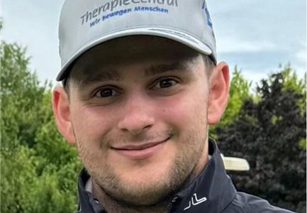 A young man with a smiling face is wearing a gray cap with the logo "TherapieCentral". In the background, trees can be seen.