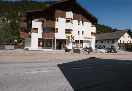 A building with wooden cladding and large windows stands on a street. Several cars are parked in the lot, and mountains can be seen in the background.