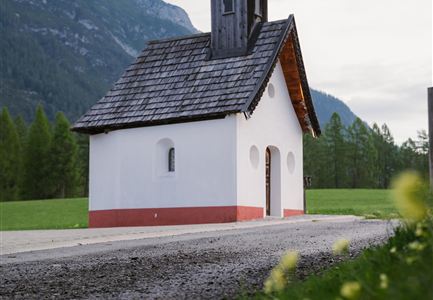 A small, picturesque chapel stands in a green meadow, surrounded by mountains. The sky is clear and the light is soft.